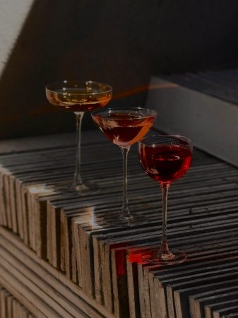 Três taças com bebidas em tons de dourado, âmbar e vermelho sobre degraus de concreto iluminados pela luz natural