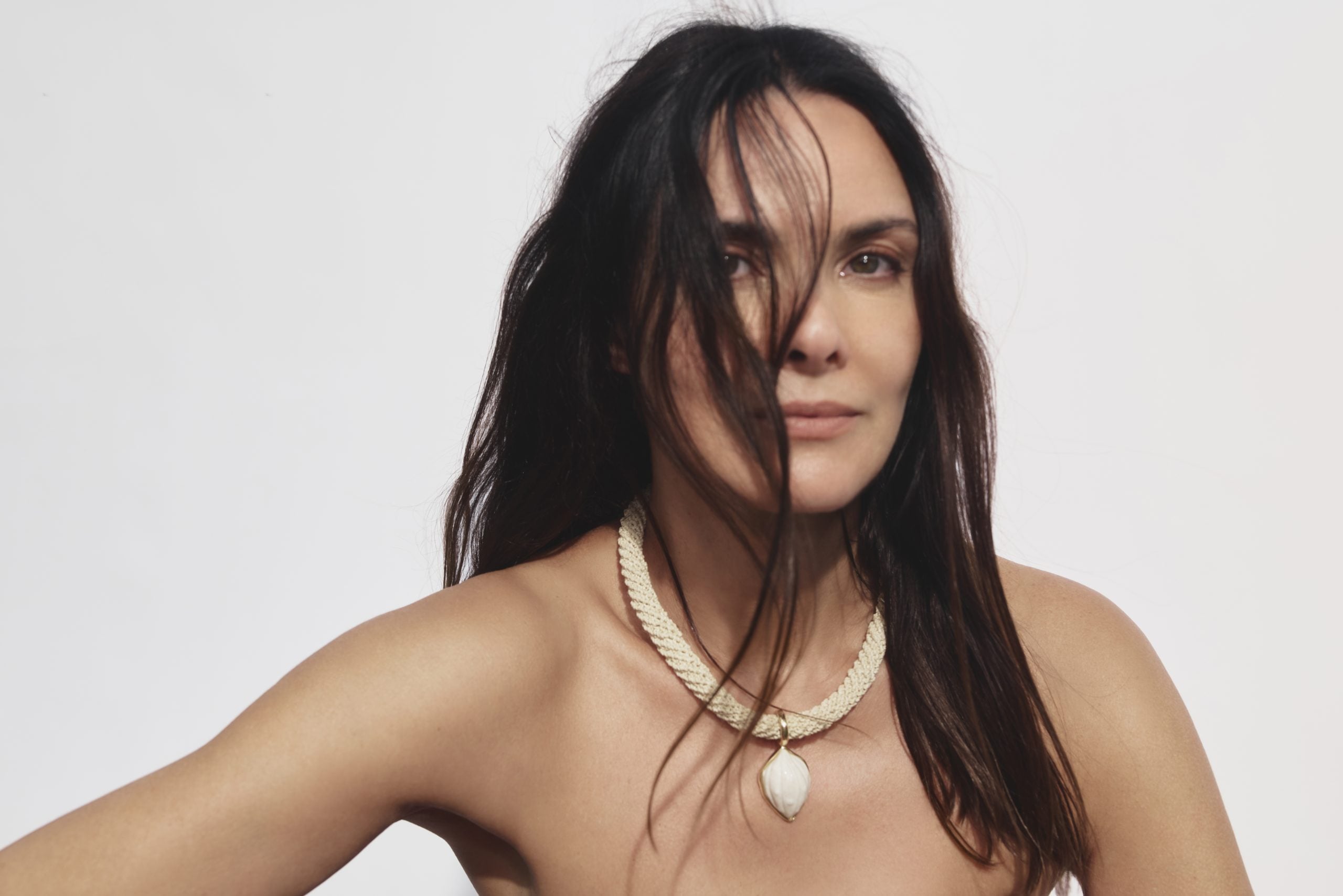 Estilista Cris Barros. Mulher de cabelos castanhos soltos posa contra fundo branco, usando colar de corda clara com pingente em formato orgânico. Cris Barros reforça relação com Curitiba.