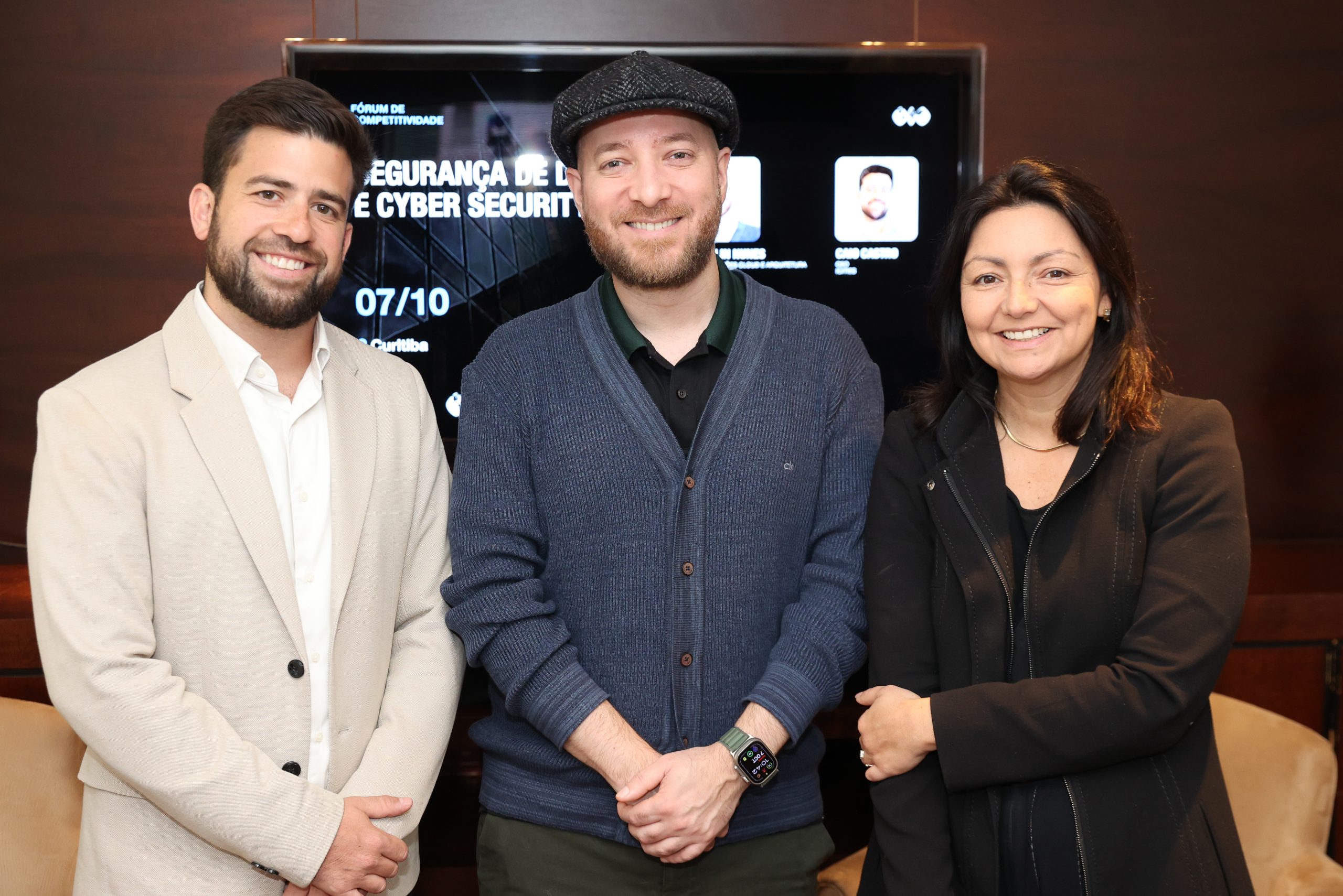 Três pessoas sorrindo, posando em frente a uma tela de evento sobre segurança de dados e cibersegurança.