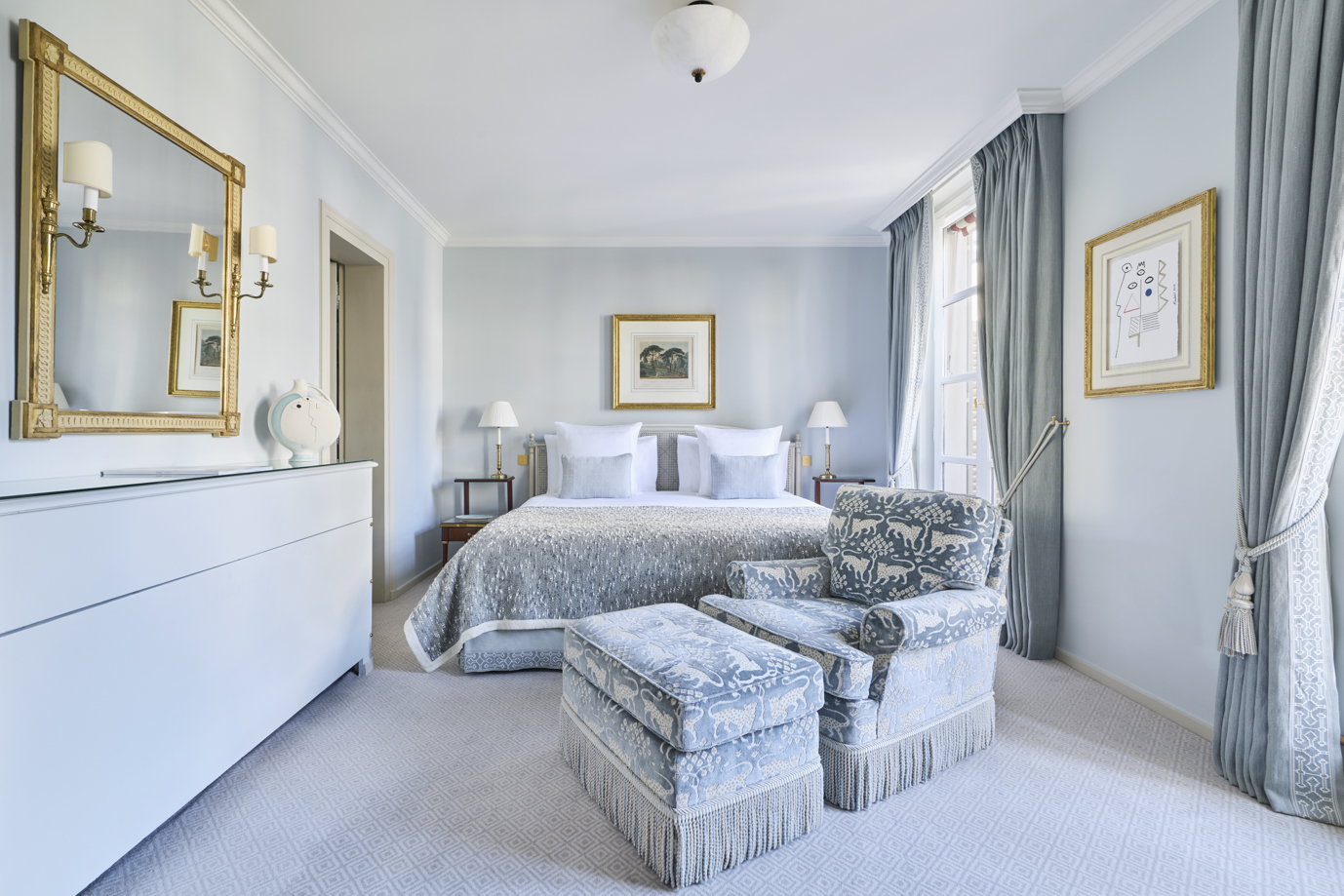 Quarto elegante em tons de azul claro e branco, com cama ampla, poltrona e puff estampados, cortinas longas e molduras douradas nas paredes.