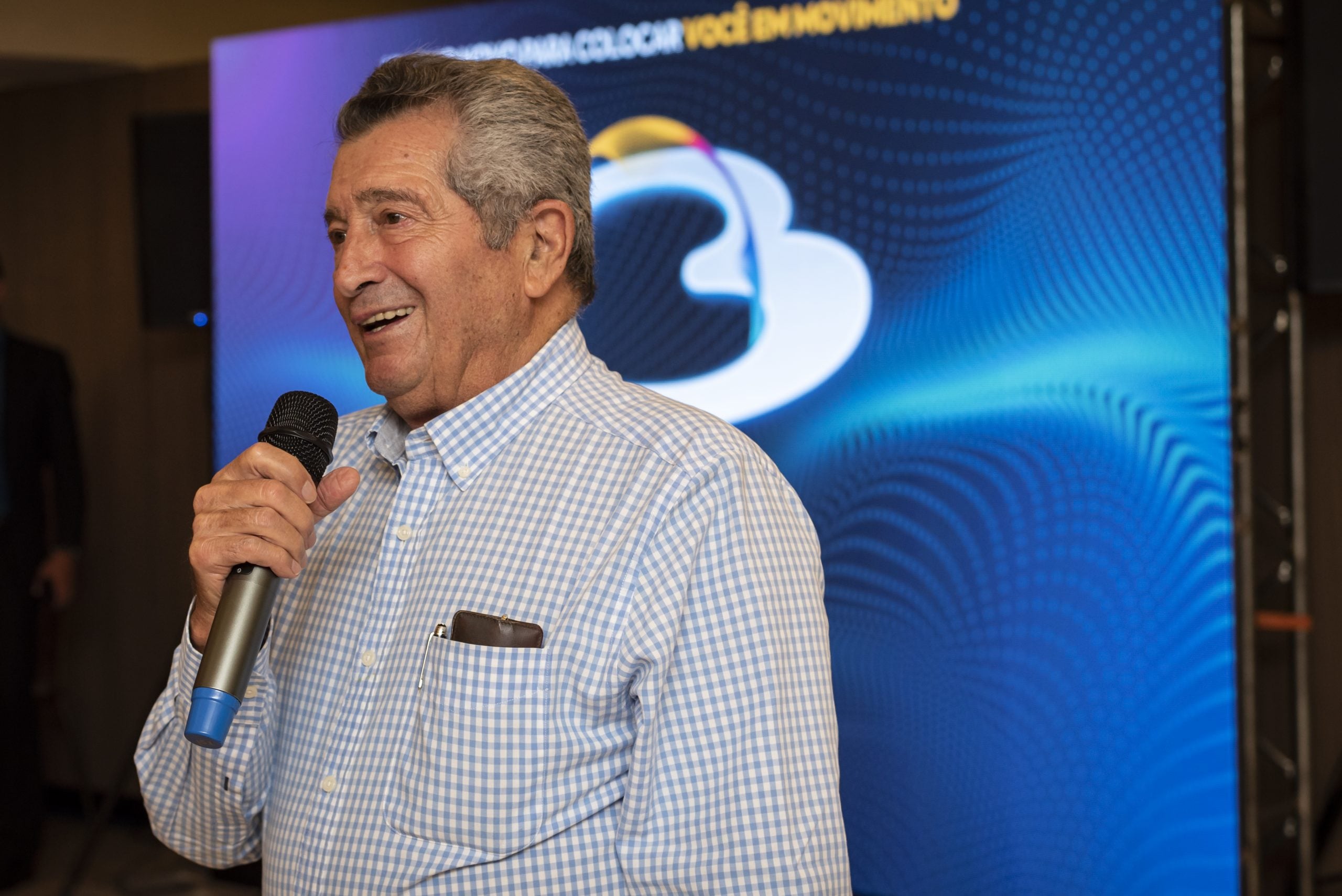 Homem sorridente segurando um microfone e falando em um evento, com fundo azul e iluminação de palco.