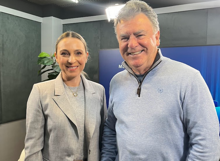 Duas pessoas sorrindo em um estúdio iluminado, vestindo roupas em tons de cinza e azul-claro.