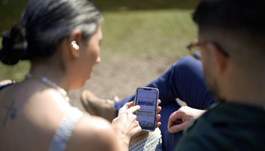 Duas pessoas sentadas ao ar livre olhando para um celular, com uma delas apontando para a tela.