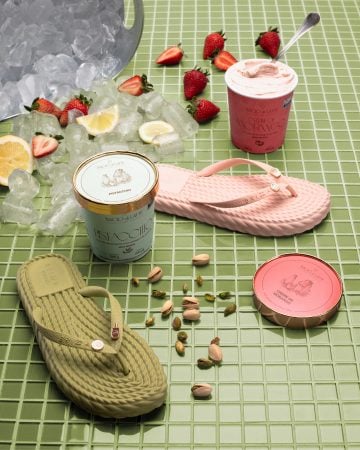 Produtos da Bacio di Latte aparecem ao lado de duas sandálias coloridas, com morangos, limões e gelo espalhados sobre uma superfície verde.