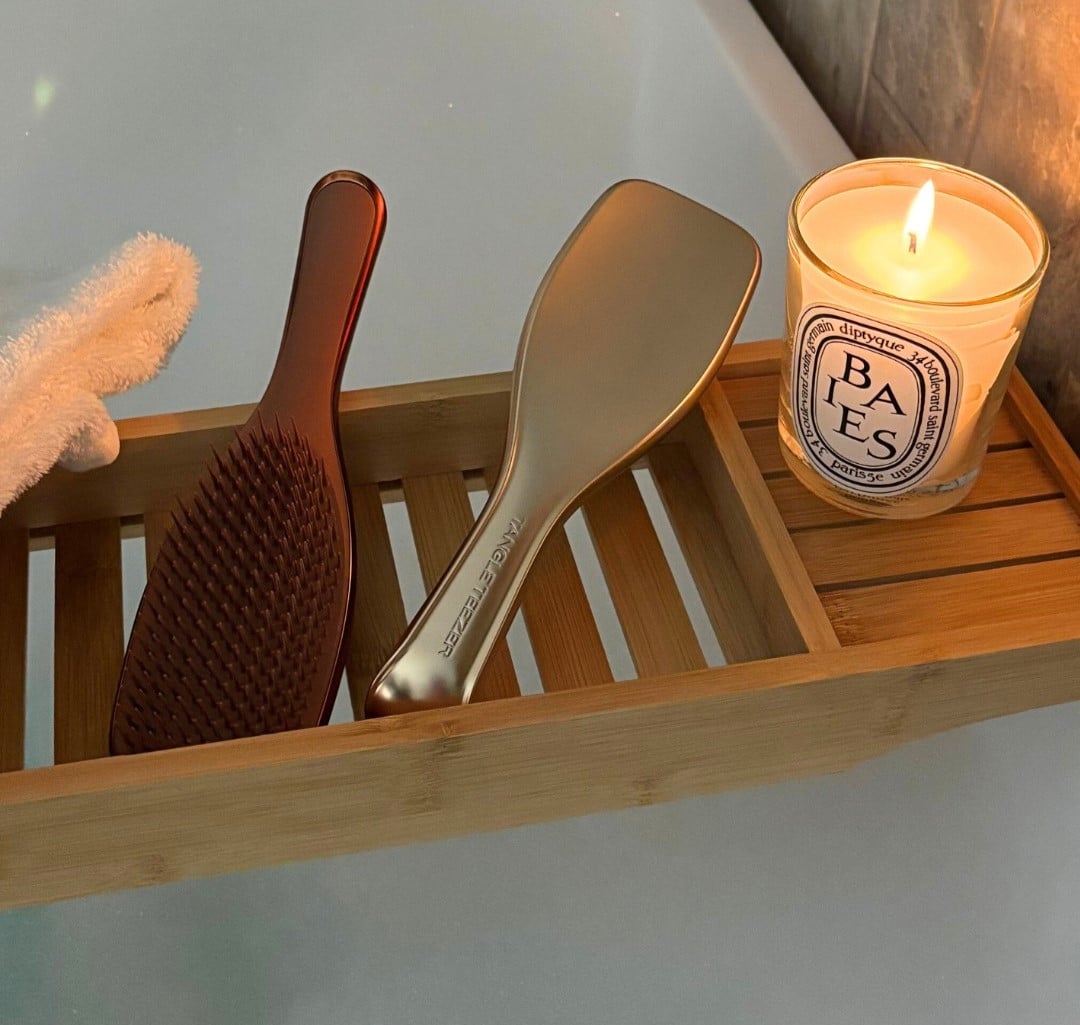 Duas escovas em tons neutros estão sobre uma bandeja de madeira ao lado de uma vela acesa, criando um clima de banho e relaxamento