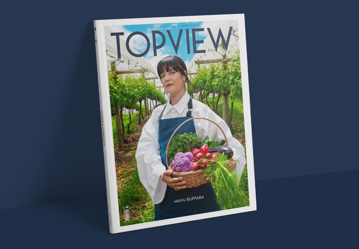 Uma mulher posa em meio a parreirais, vestindo avental azul e camisa branca, segurando uma cesta cheia de vegetais coloridos como couve-flor roxa, rabanetes e ervas frescas. A imagem compõe a capa da revista TOPVIEW, destacando a atmosfera natural e a conexão com ingredientes locais.