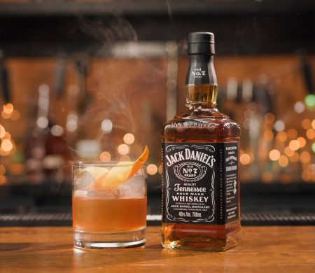 A foto mostra uma garrafa de Jack Daniel’s ao lado de um coquetel com gelo e casca de laranja sobre um balcão de bar iluminado por luzes suaves ao fundo.