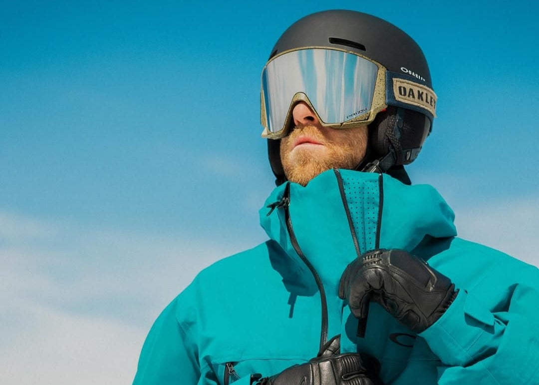 Pessoa com roupa de esqui azul, capacete preto e óculos espelhados, olhando para o horizonte sob um céu azul claro.