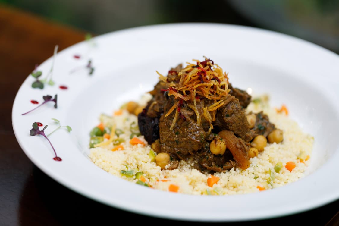 A foto mostra um prato de inspiração marroquina, com carne cozida lentamente servida sobre cuscuz com legumes e grãos-de-bico. A montagem é detalhada, com fios crocantes por cima e decoração delicada na borda do prato.
