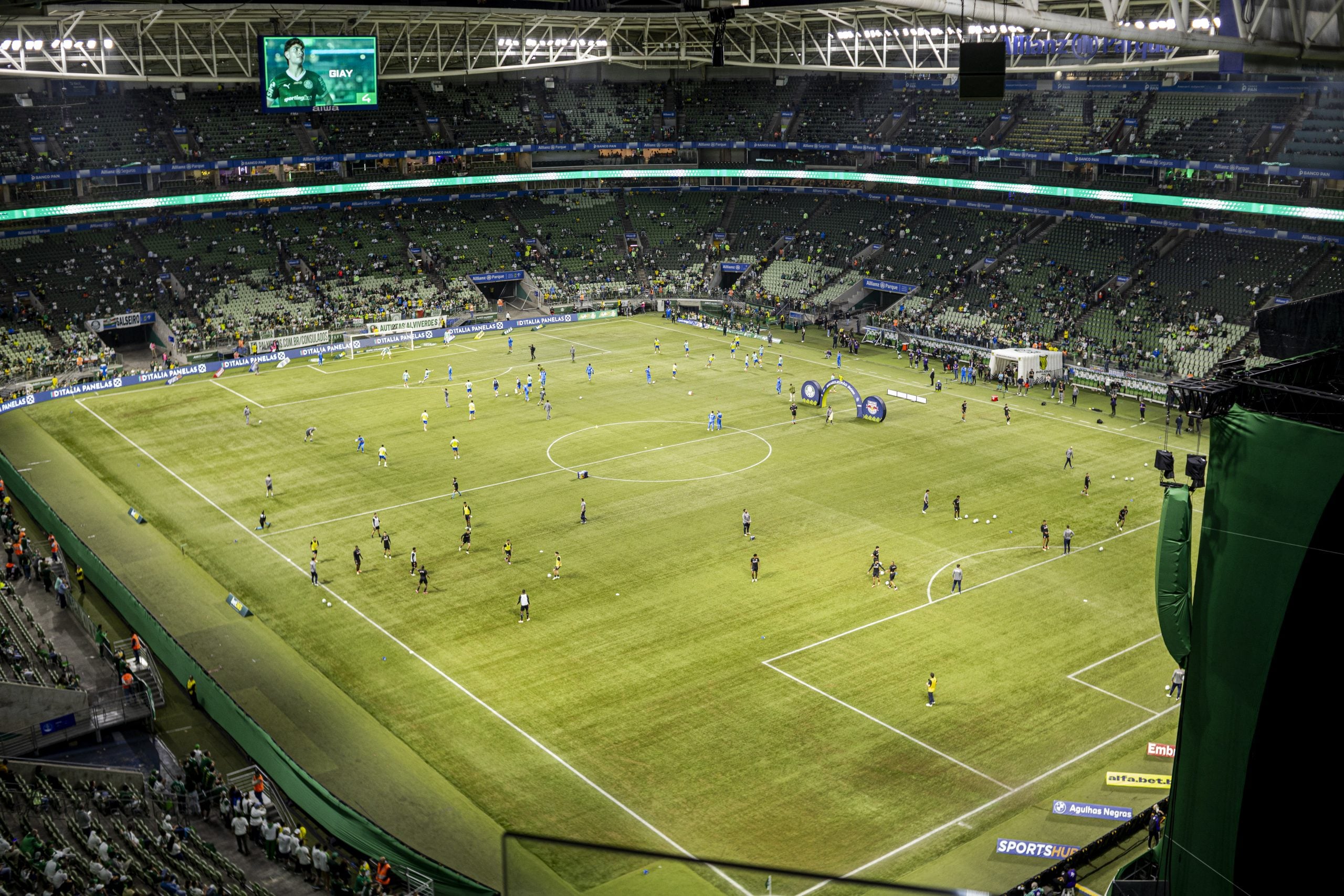 A imagem mostra um estádio de futebol iluminado, com jogadores realizando o aquecimento em campo e torcedores ocupando parcialmente as arquibancadas antes do início de uma partida. Backstage Mirante, Allianz Parque.