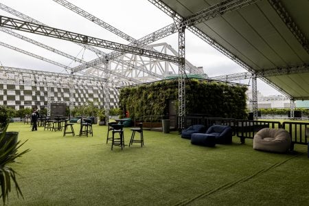 A imagem mostra um espaço ao ar livre coberto, com grama sintética, mesas altas, pufes e uma estrutura moderna com paredes verdes e vista para um estádio. Backstage Mirante, Allianz Parque.