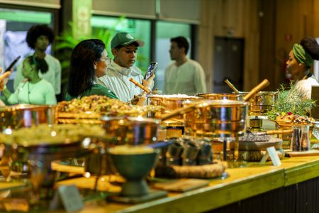 A imagem mostra um buffet elegante com diversas comidas em panelas de cobre, enquanto pessoas observam e tiram fotos do ambiente no Backstage Mirante, no Allianz Parque.