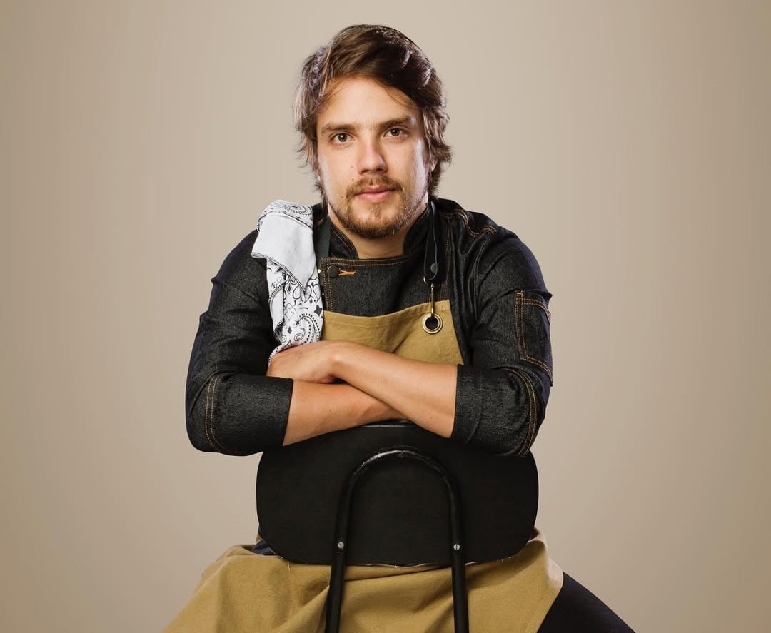 home com barba, com roupa de chef de cozinha, com braços cruzados e sentado em uma cadeira