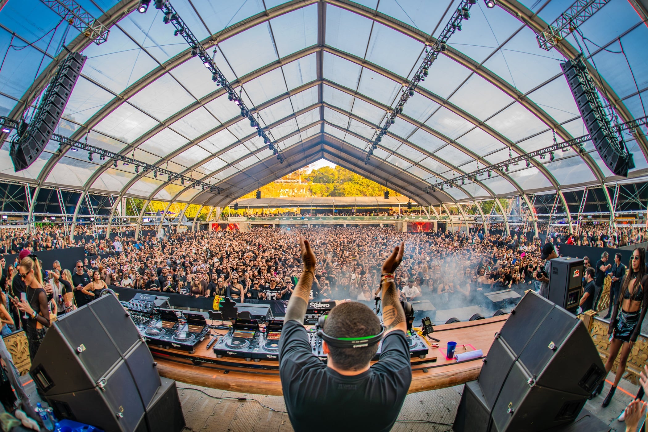 A imagem mostra um DJ de costas diante de uma multidão animada em um festival sob uma tenda transparente, com luz natural realçando a atmosfera vibrante do evento.
