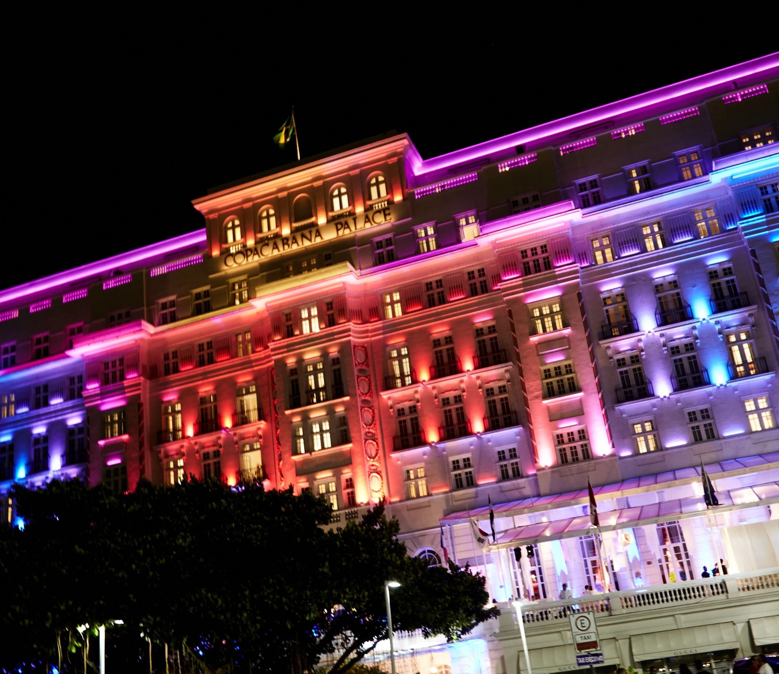 A imagem mostra a fachada do Copacabana Palace iluminada em tons vibrantes à noite, com pessoas circulando na entrada e clima festivo ao redor.