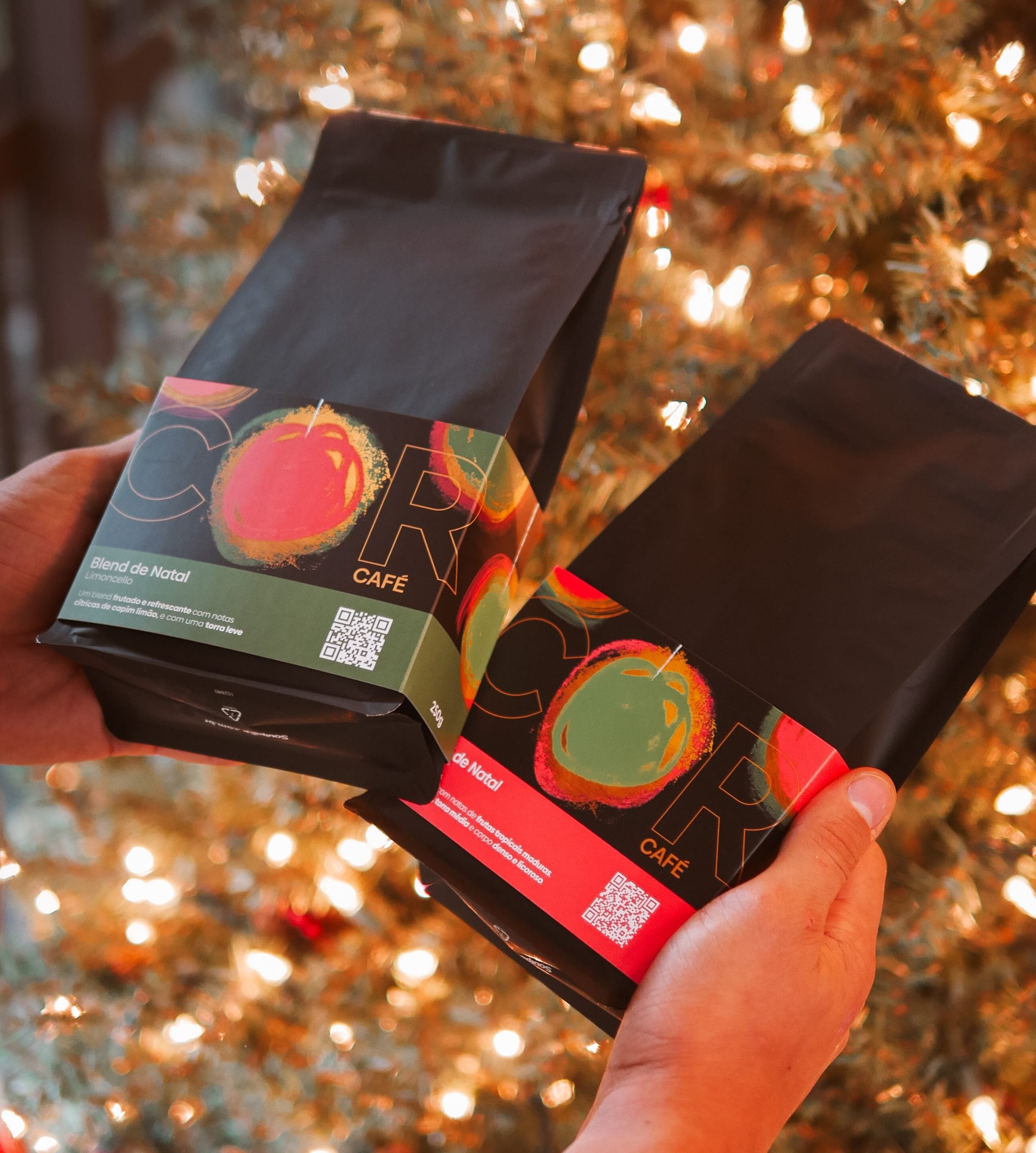 Mãos seguram dois pacotes dos Blends de Natal do COR Café diante de uma árvore iluminada, destacando os rótulos coloridos e festivos que reforçam o clima natalino da coleção.