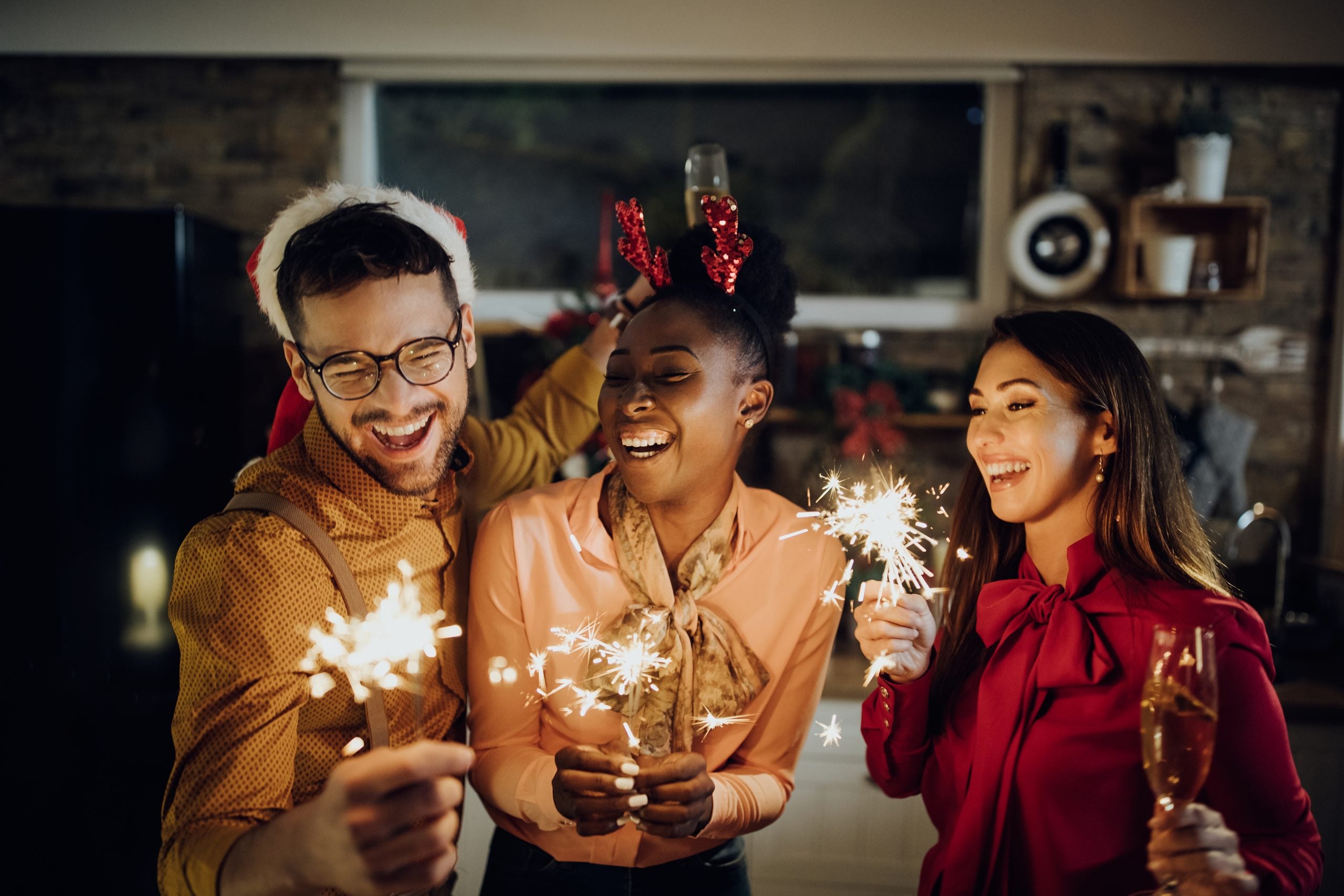 A imagem mostra três pessoas comemorando com velas estrelinhas, brindando e sorrindo em um ambiente decorado para o fim de ano. Elas usam acessórios festivos e transmitem um clima alegre e descontraído.