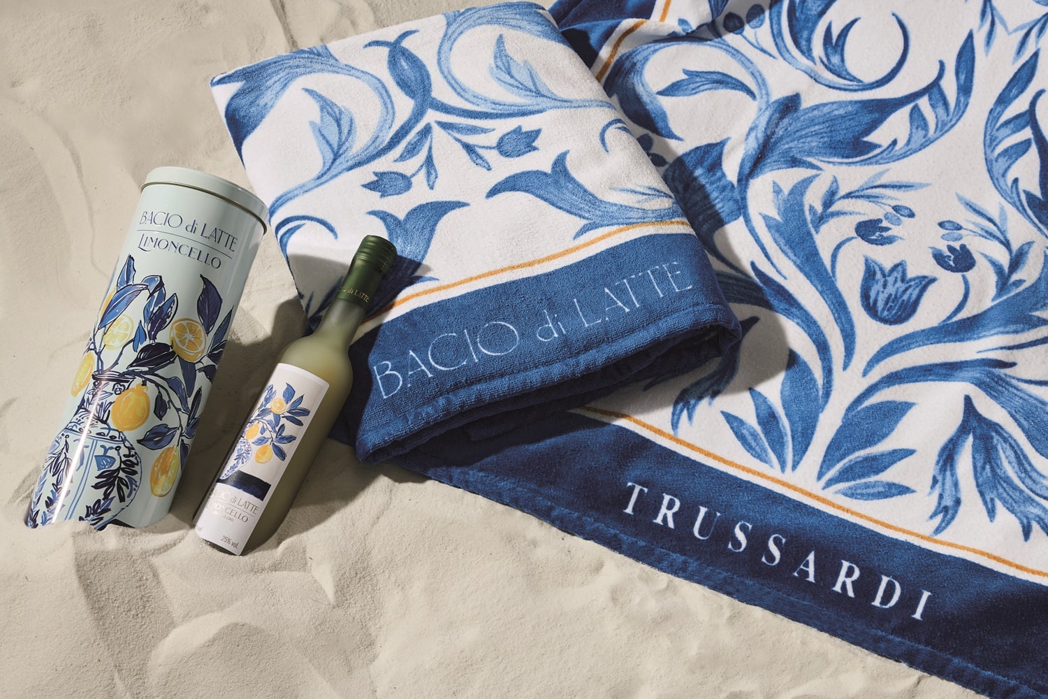 A imagem mostra um conjunto elegante na areia, com toalhas estampadas em azul e branco das marcas Bacio di Latte e Trussardi, exibindo um design floral sofisticado. Ao lado, há uma lata e uma garrafa de Limoncello, ambas decoradas com ilustrações de limões, criando uma atmosfera fresca e mediterrânea.